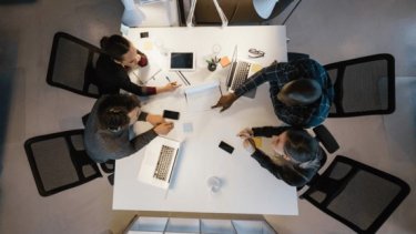 A group of diverse individuals seated around a table, each using a laptop, engaged in discussion or collaboration.
